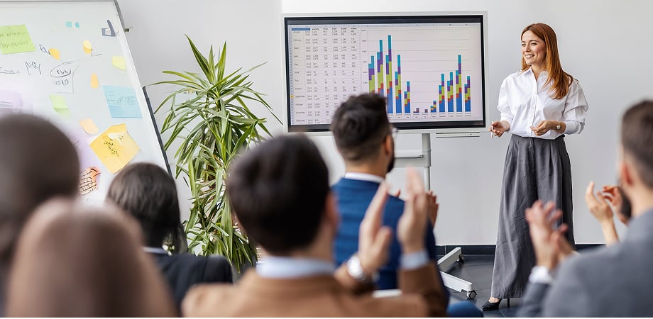 a woman delivering a presentation in front of a large crowd using graphs displayed on a wall-mounted screen