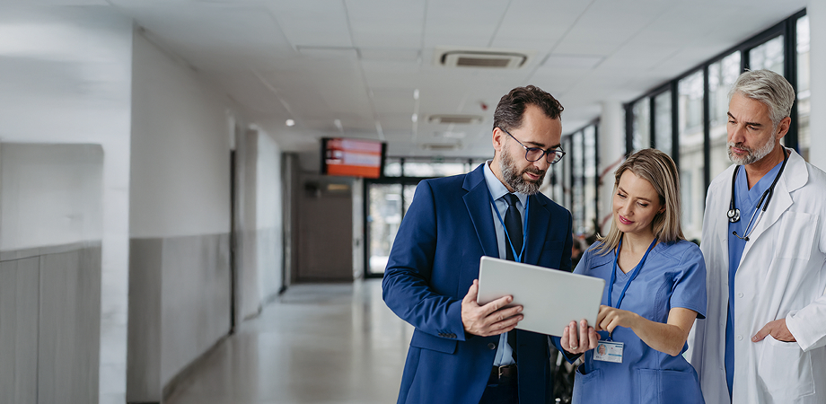 a man in a blue suit having a discussion with two healthcare professionals while looking at a tablet screen
