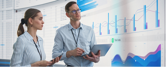 a man and a woman having a discussion while looking at a wall of screens displaying charts and graphs