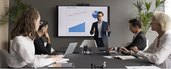a man delivering a presentation to colleagues in a conference room while standing in front of a large screen displaying charts and graphs