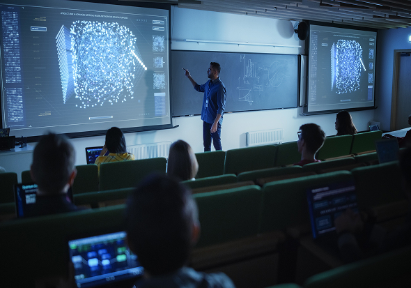 a man delivering a lecture in a modern classroom while standing in front of a blackboard flanked by two large screens